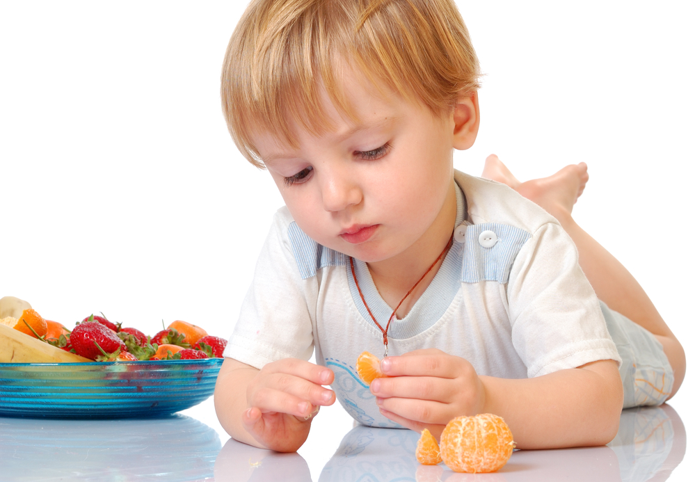 Ребенок трёх лет в белой футболке, лёжа на животе, держит в руках мандарин